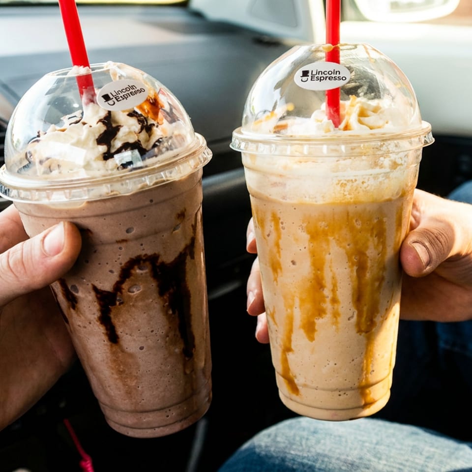 Two hands hold Lincoln Espresso chocolate and coffee blended drinks with whipped cream inside a car.