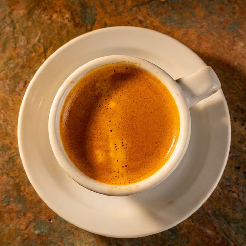 A white cup of espresso with rich crema sits on a saucer on a textured brown table.